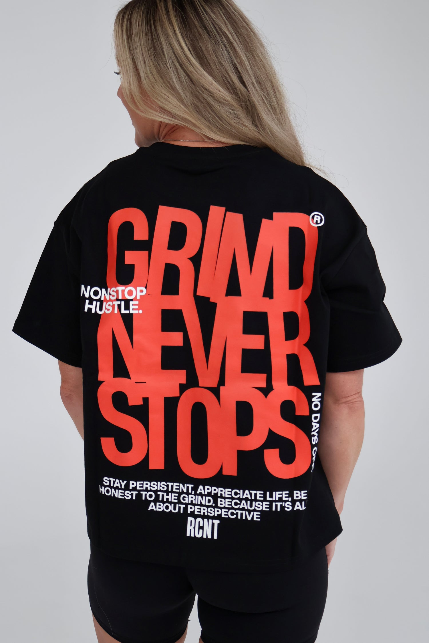 Woman wearing black oversized t-shirt with bold red 'GRIND NEVER STOPS' and motivational text on back