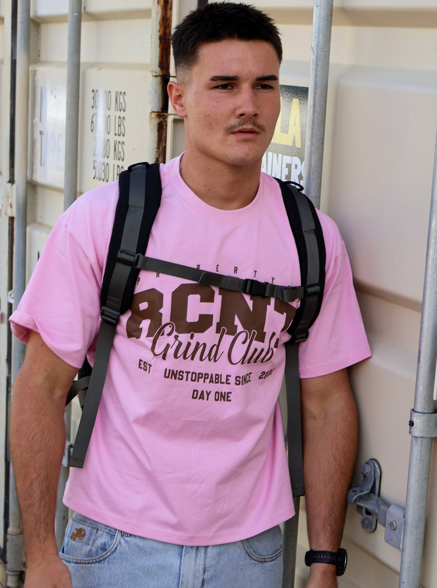 Man wearing pink RCNT Grind Club graphic t-shirt, backpack, and jeans standing outdoors