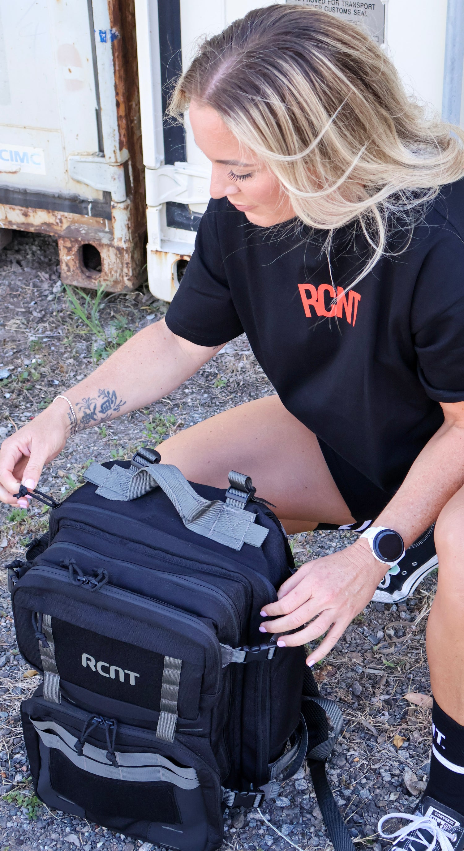 Person wearing RCNT t-shirt crouching outdoors, adjusting black RCNT tactical backpack