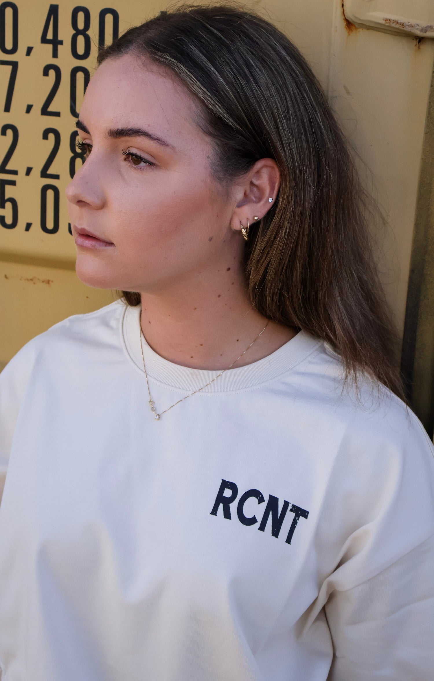 Woman wearing white RCNT graphic t-shirt, gold earrings and necklace, in urban outdoor setting