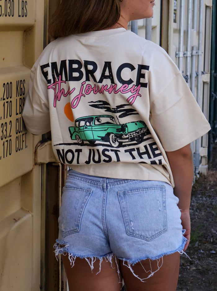 Woman in light denim shorts and graphic t-shirt that reads 'Embrace the Journey,' outdoors