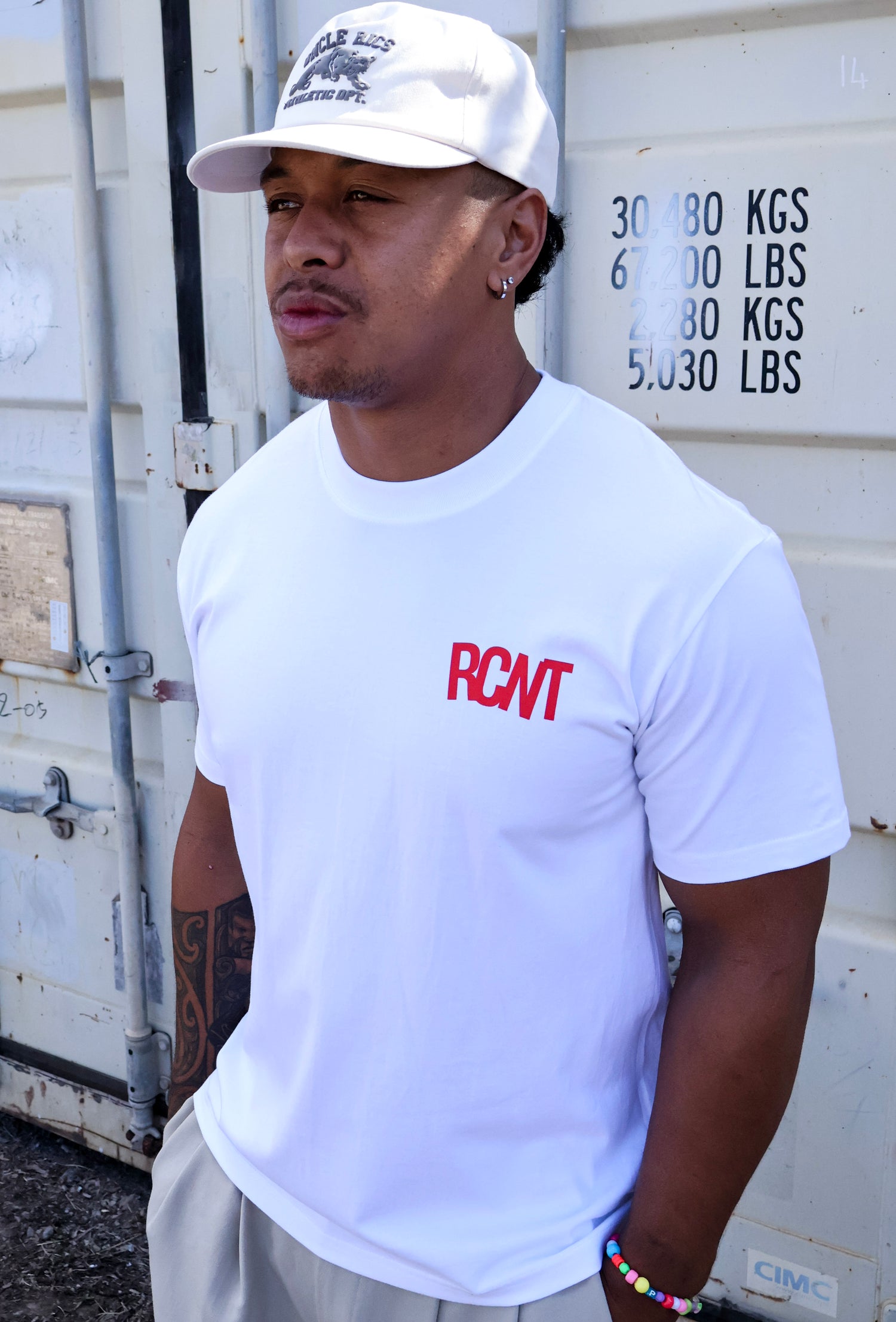 Man in white RCNT tee and cap stands by shipping container, showcasing casual streetwear style.