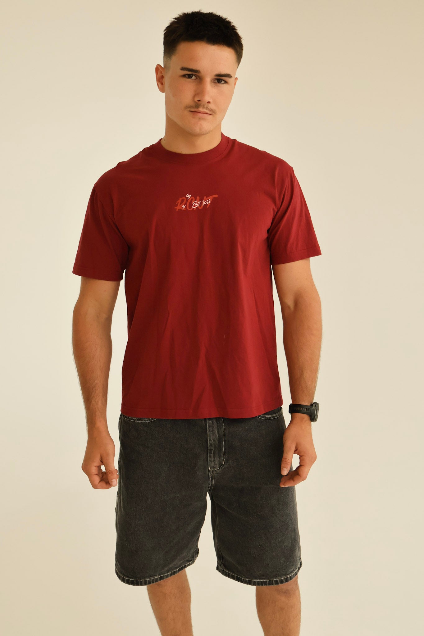 Man in red graphic t-shirt and black denim shorts standing against neutral background