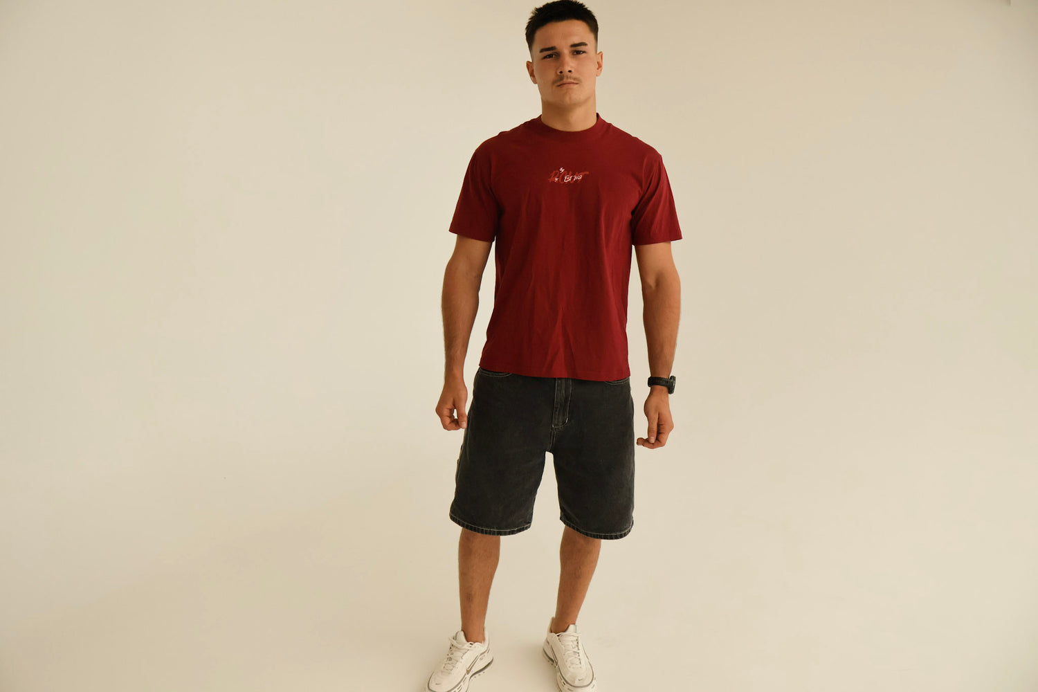 Man wearing a red graphic t-shirt, black denim shorts, and white sneakers in studio