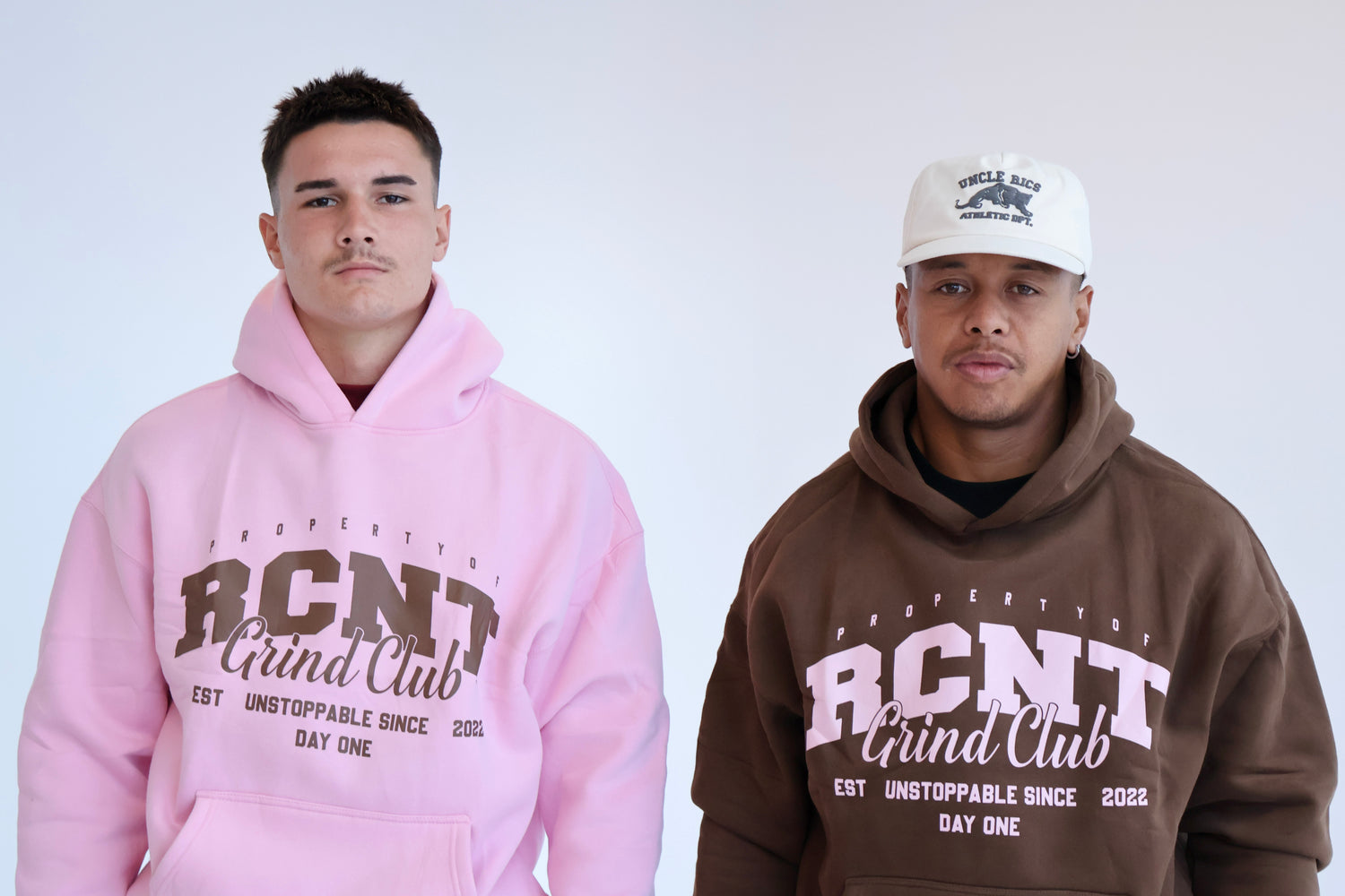 Two men modeling RCNT Grind Club hoodies, one in pink, one in brown, on a white background.