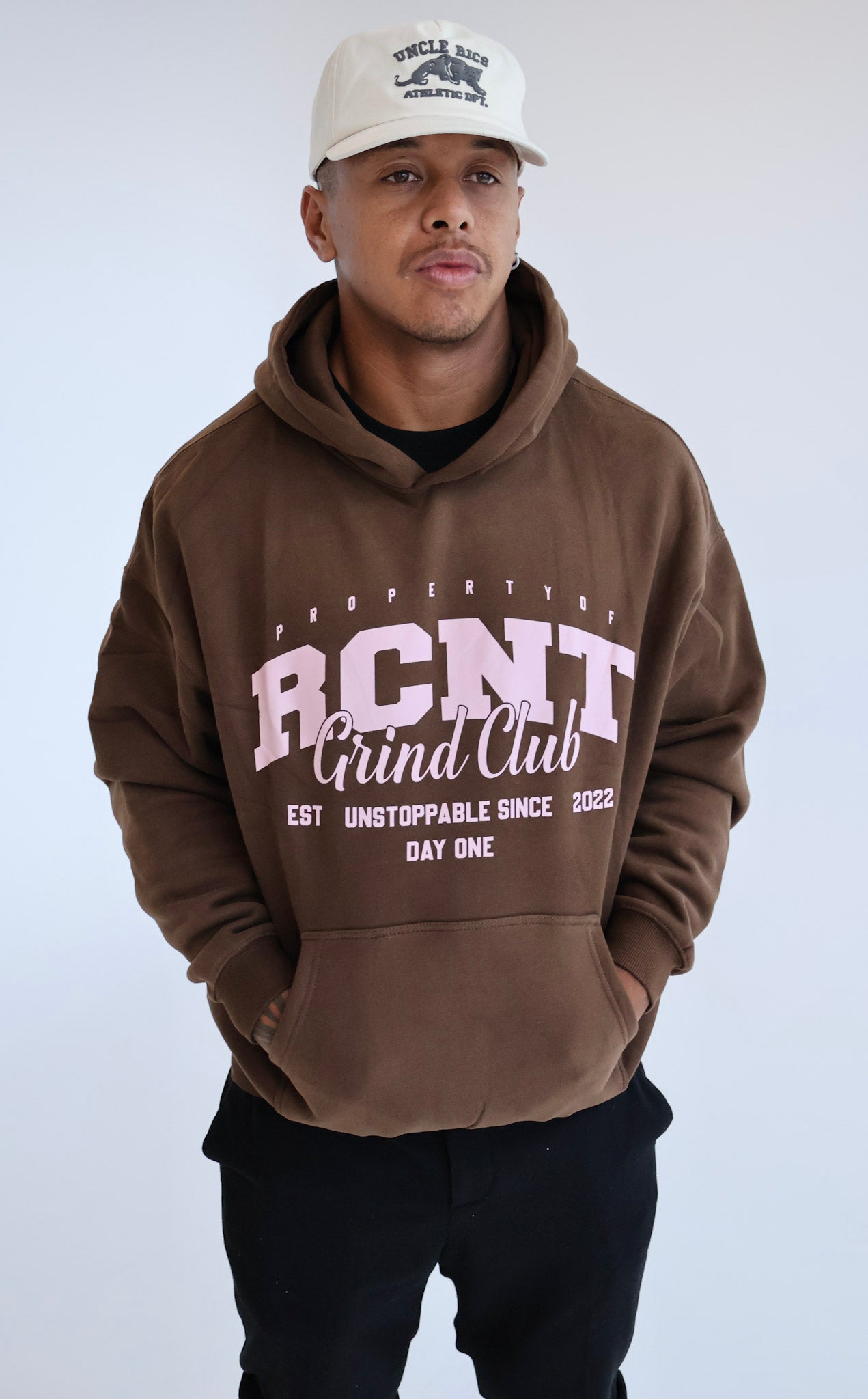 Man wearing brown RCNT Grind Club hoodie and white Uncle Ricc cap, casual streetwear style