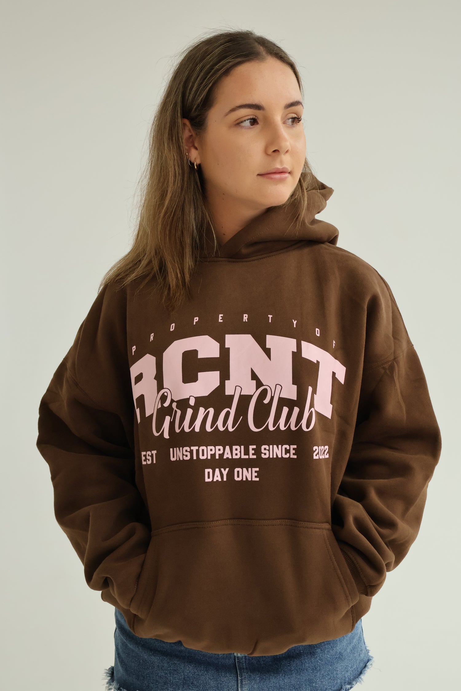 Woman in brown RCNT Grind Club hoodie with pink lettering, casual streetwear style