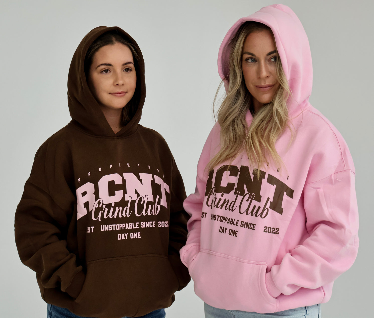 Two women wearing oversized Grind Club hoodies, one in brown and one in pink, with bold RCNT logo.