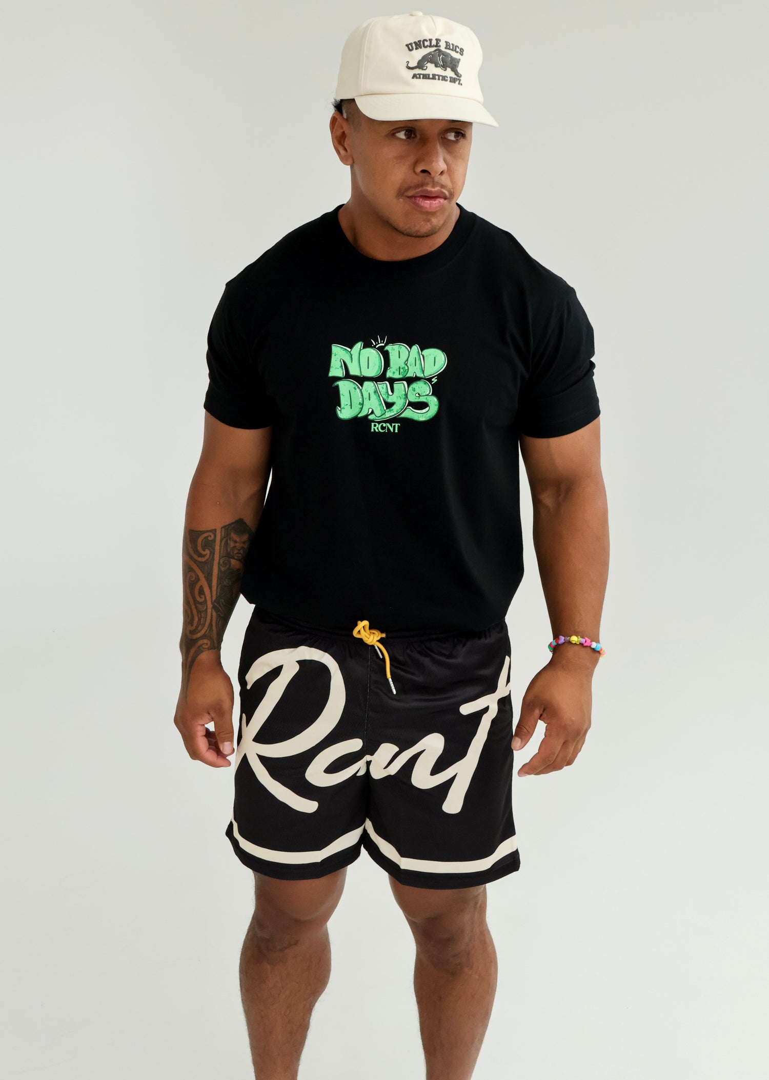 Man in black 'No Bad Days' graphic t-shirt, logo shorts, and cream athletic cap standing indoors