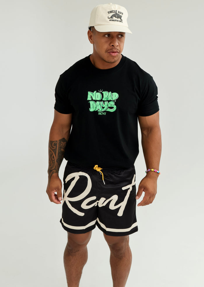 Man in black 'No Bad Days' graphic t-shirt, logo shorts, and cream athletic cap standing indoors