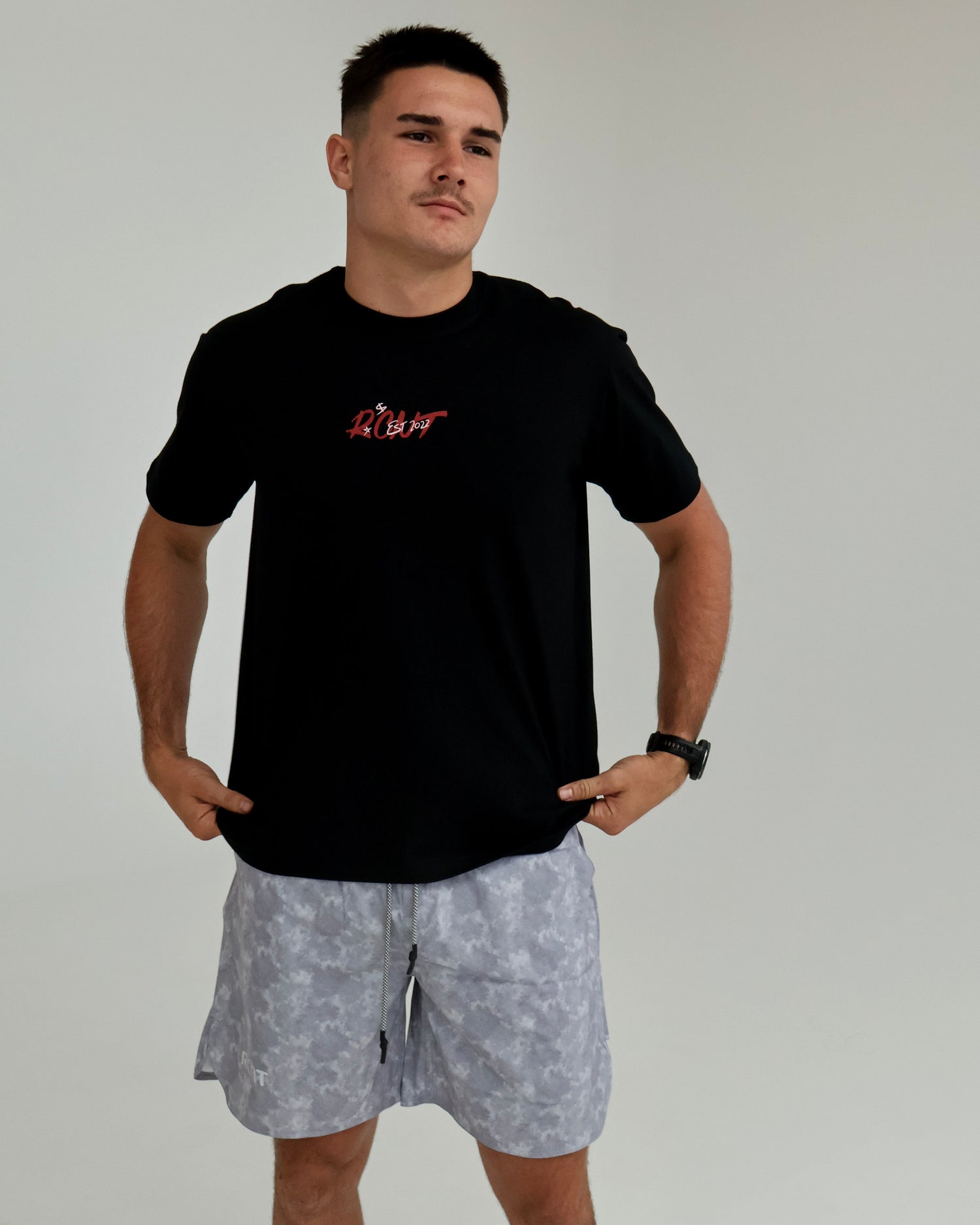 Man wearing a black graphic t-shirt and light grey camo shorts in minimal indoor setting