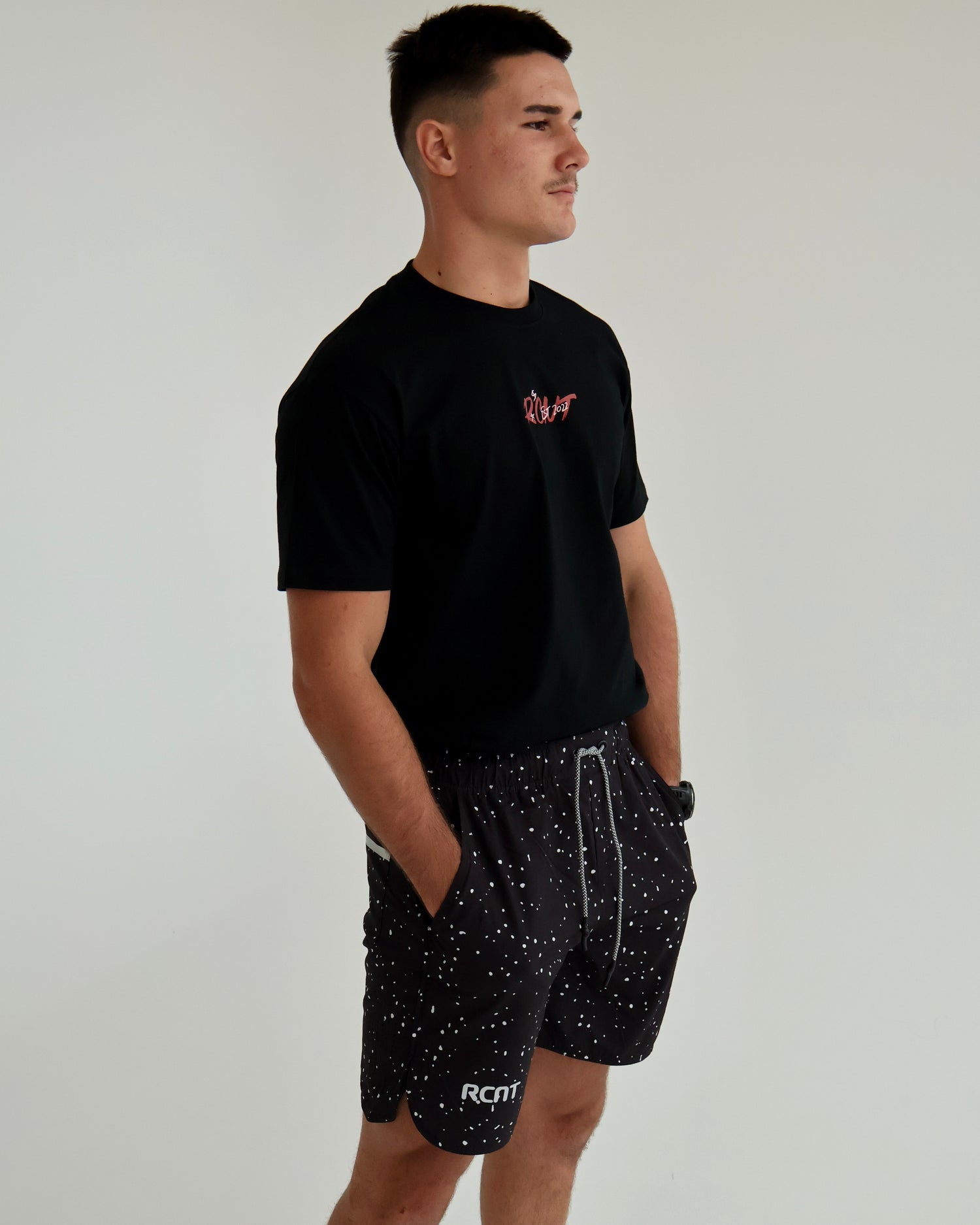 Man wearing black graphic t-shirt and black spotted shorts with RCNT logo, minimal background