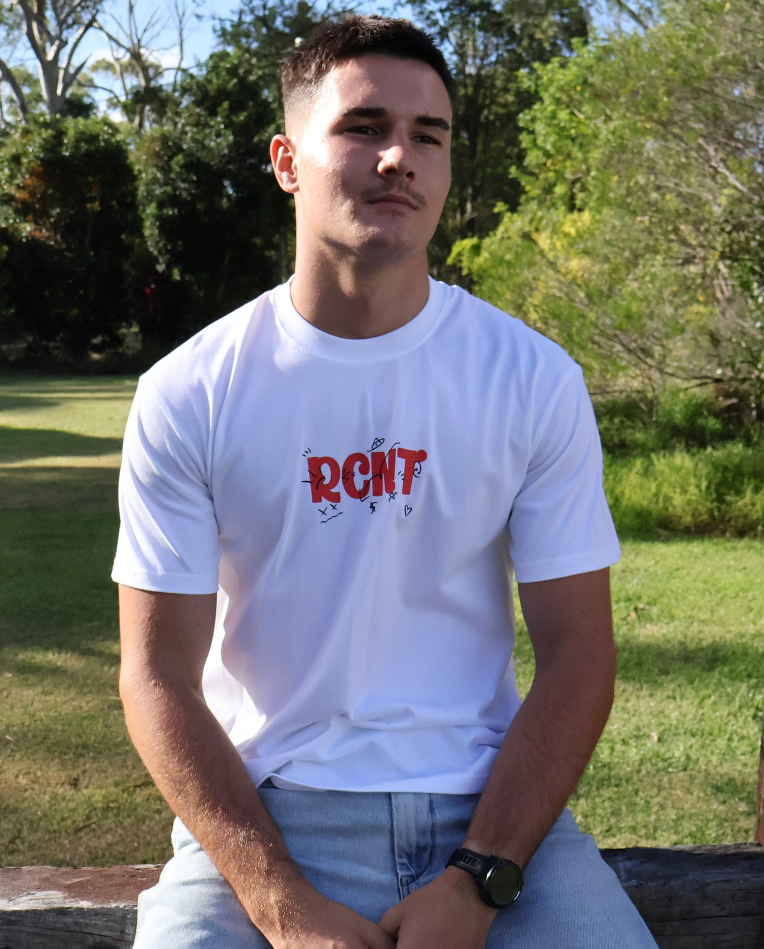 Man wearing white graphic RCNT t-shirt and jeans sitting outdoors in sunny park