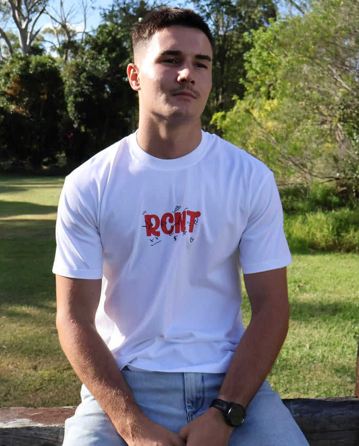 Man wearing white graphic RCNT t-shirt and jeans sitting outdoors in sunny park