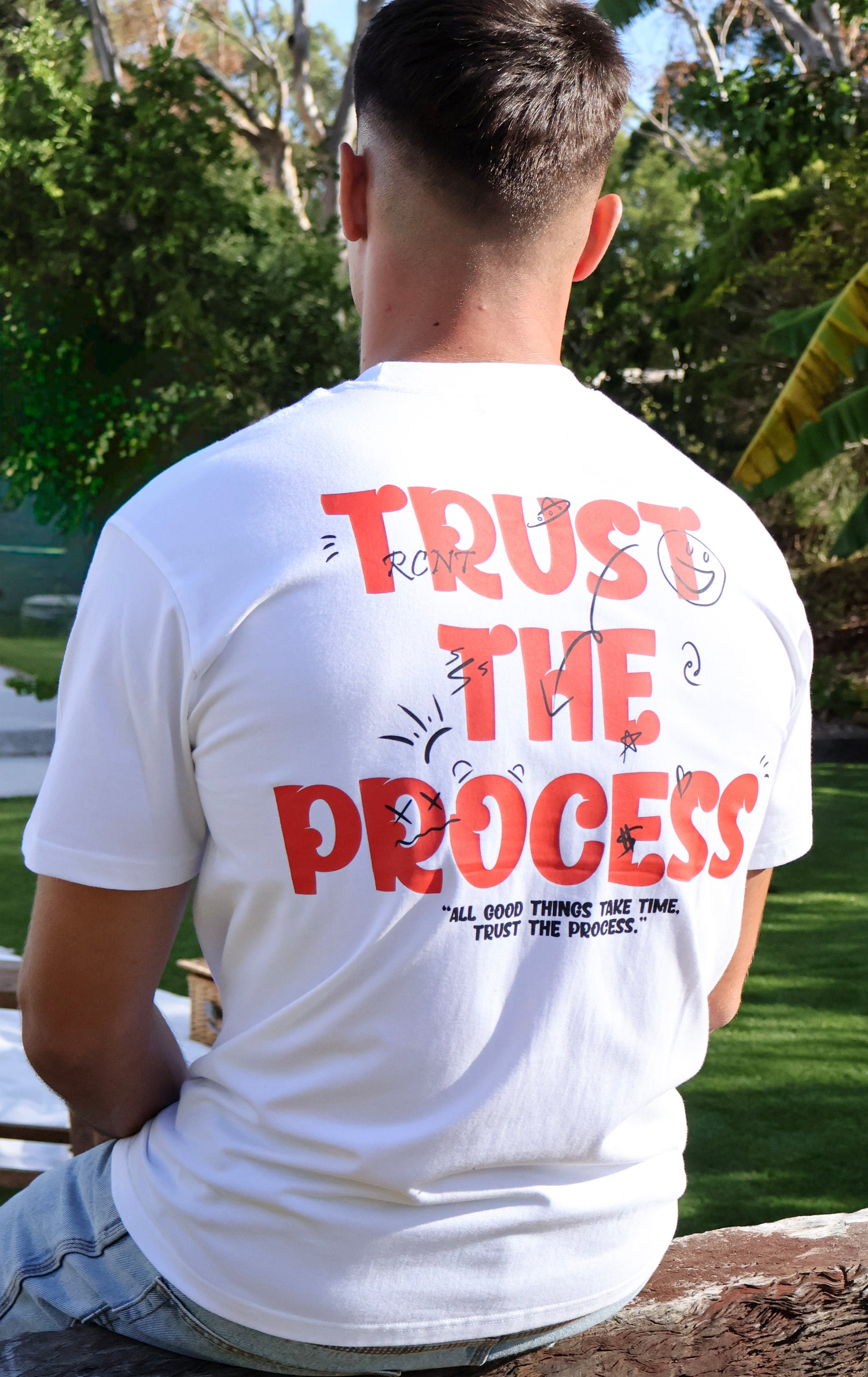 Man outdoors in white graphic t-shirt with 'Trust the Process' lettering on the back.
