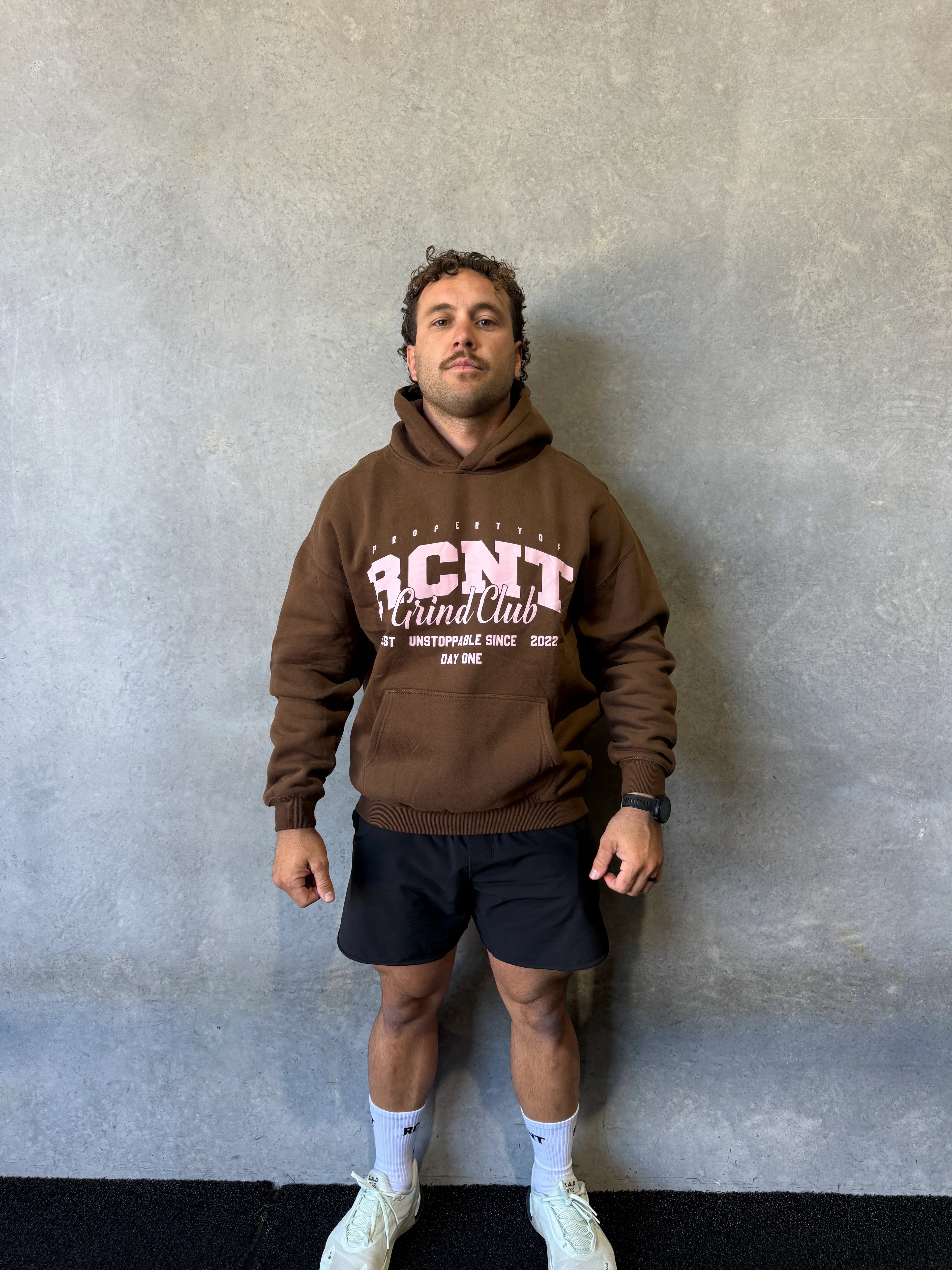 Man wearing brown hoodie with BCNT Grind Club text, black shorts, white socks, and sneakers standing by concrete wall