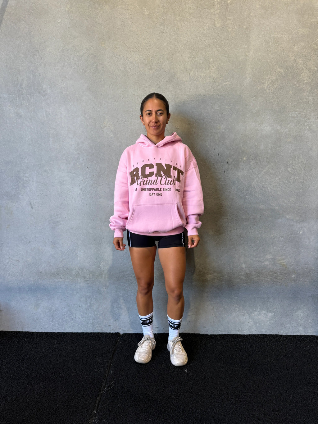 Woman in pink hoodie, black shorts, and white sneakers standing against gray wall