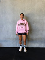 Woman in pink hoodie, black shorts, and white sneakers standing against gray wall