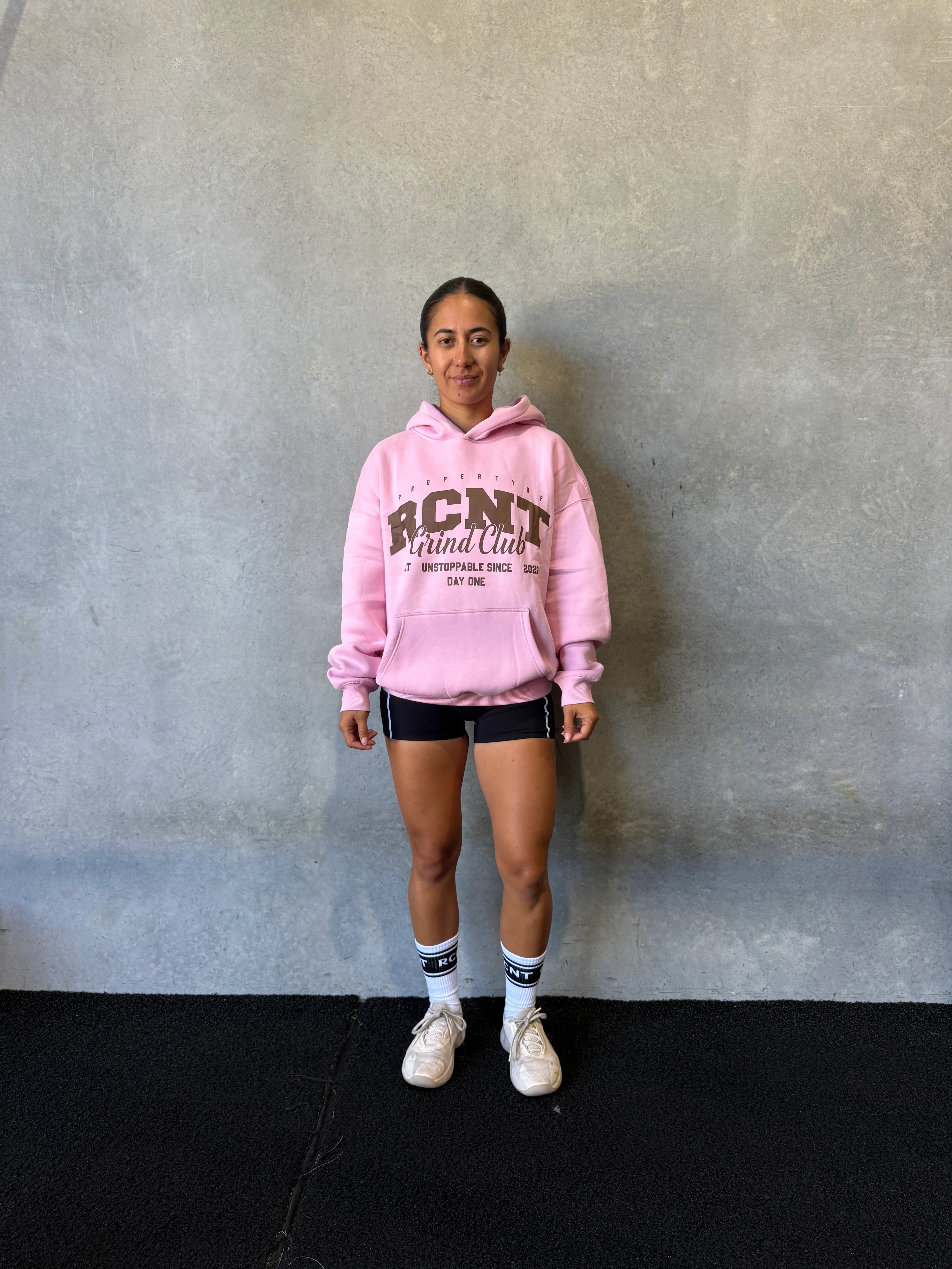 Woman in pink hoodie, black shorts, and white sneakers standing against gray wall
