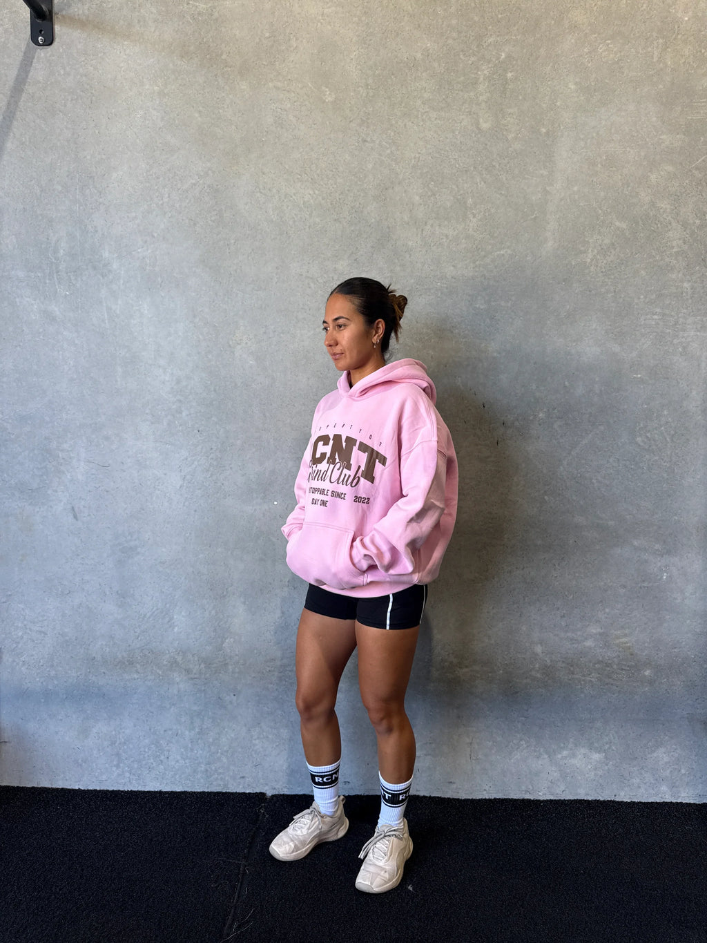 Woman in pink CNT hoodie, black shorts, white socks and sneakers standing against concrete wall