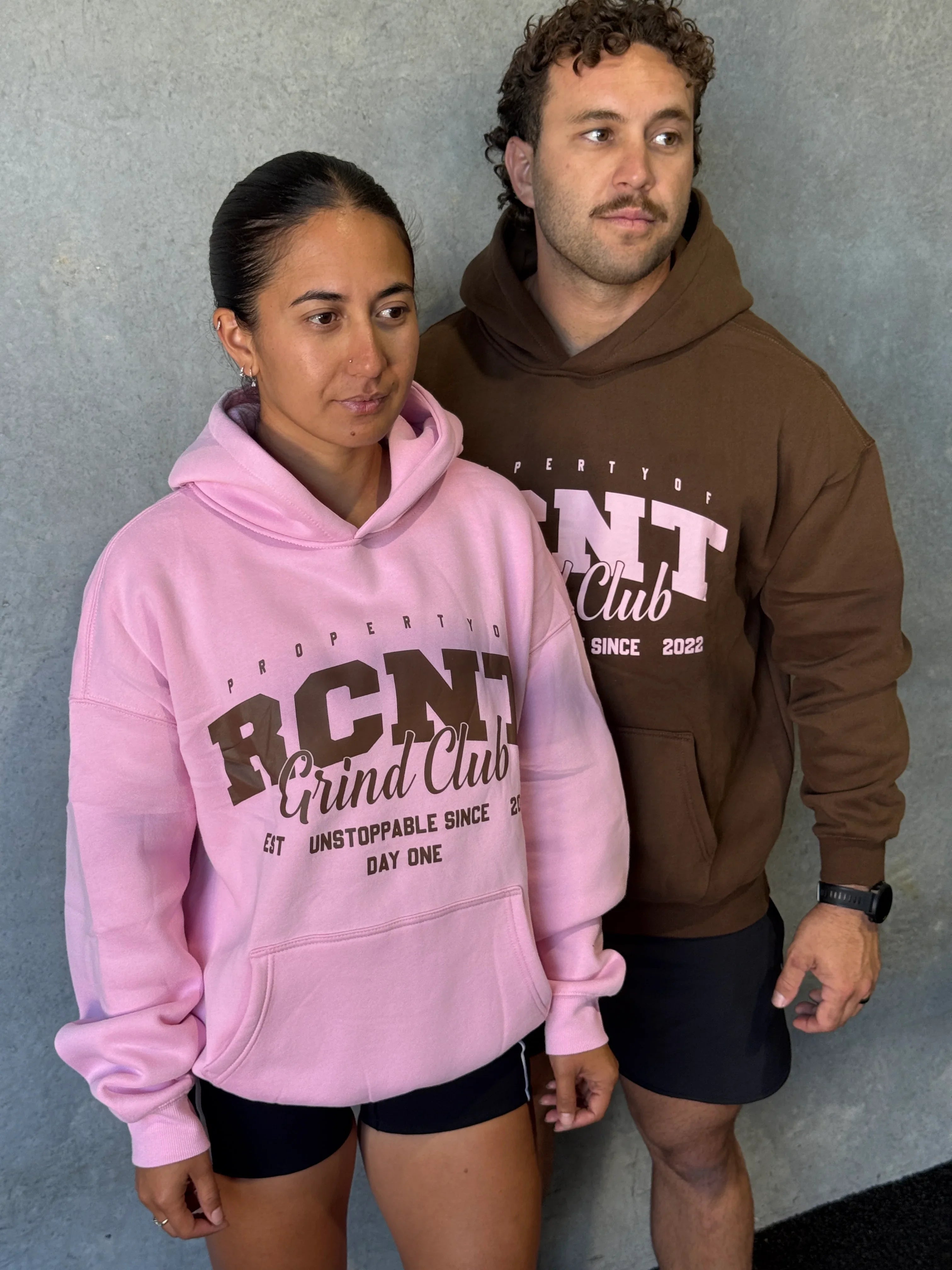 Two models wearing RCNT Grind Club hoodies in pink and brown against a gray wall