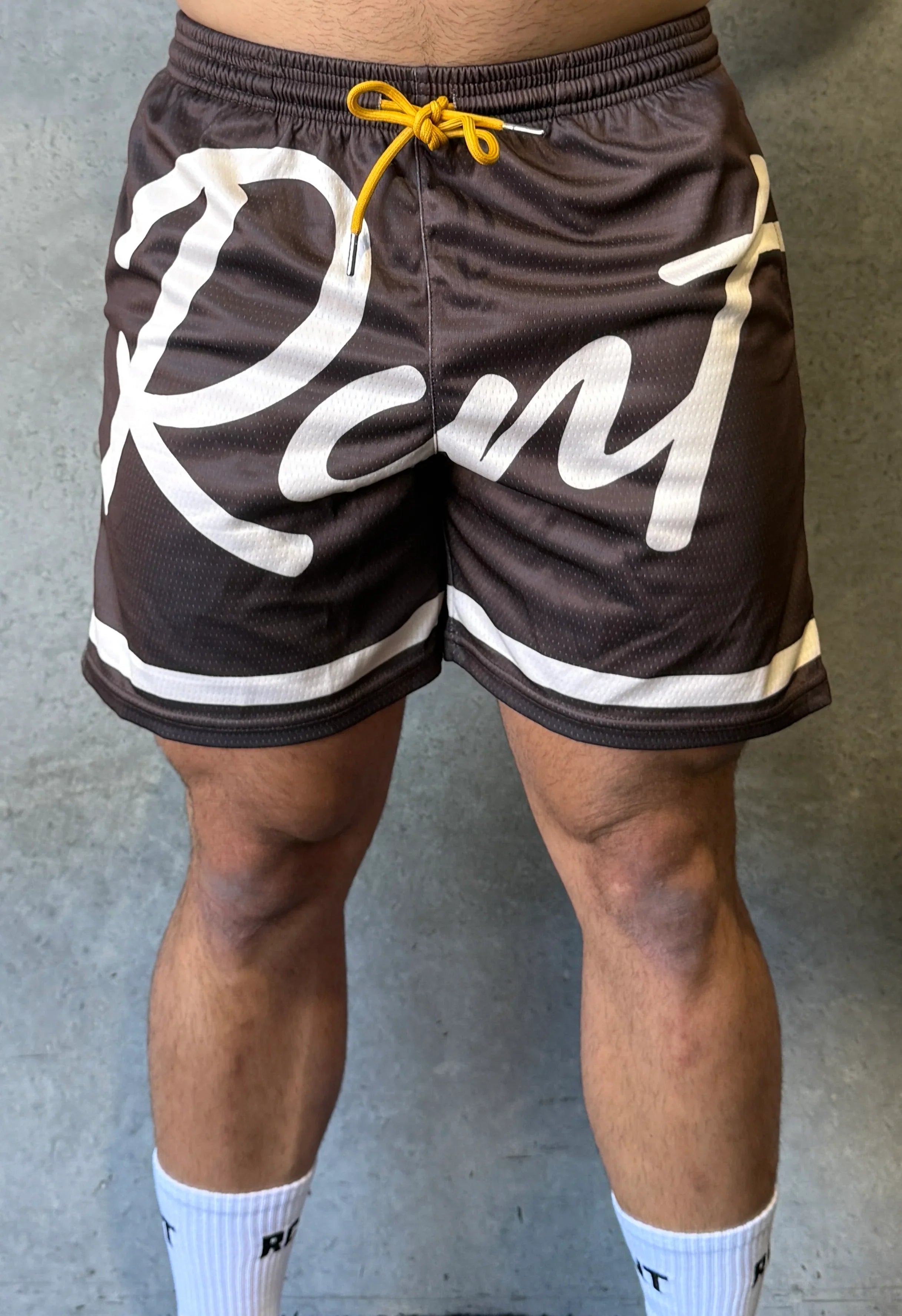 Close-up of a person wearing brown sports shorts with white 'Rant' text and yellow drawstring against gray background