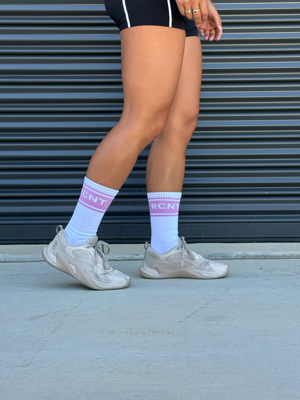 person wearing white athletic socks with pink RCNT text and beige sneakers against black corrugated wall