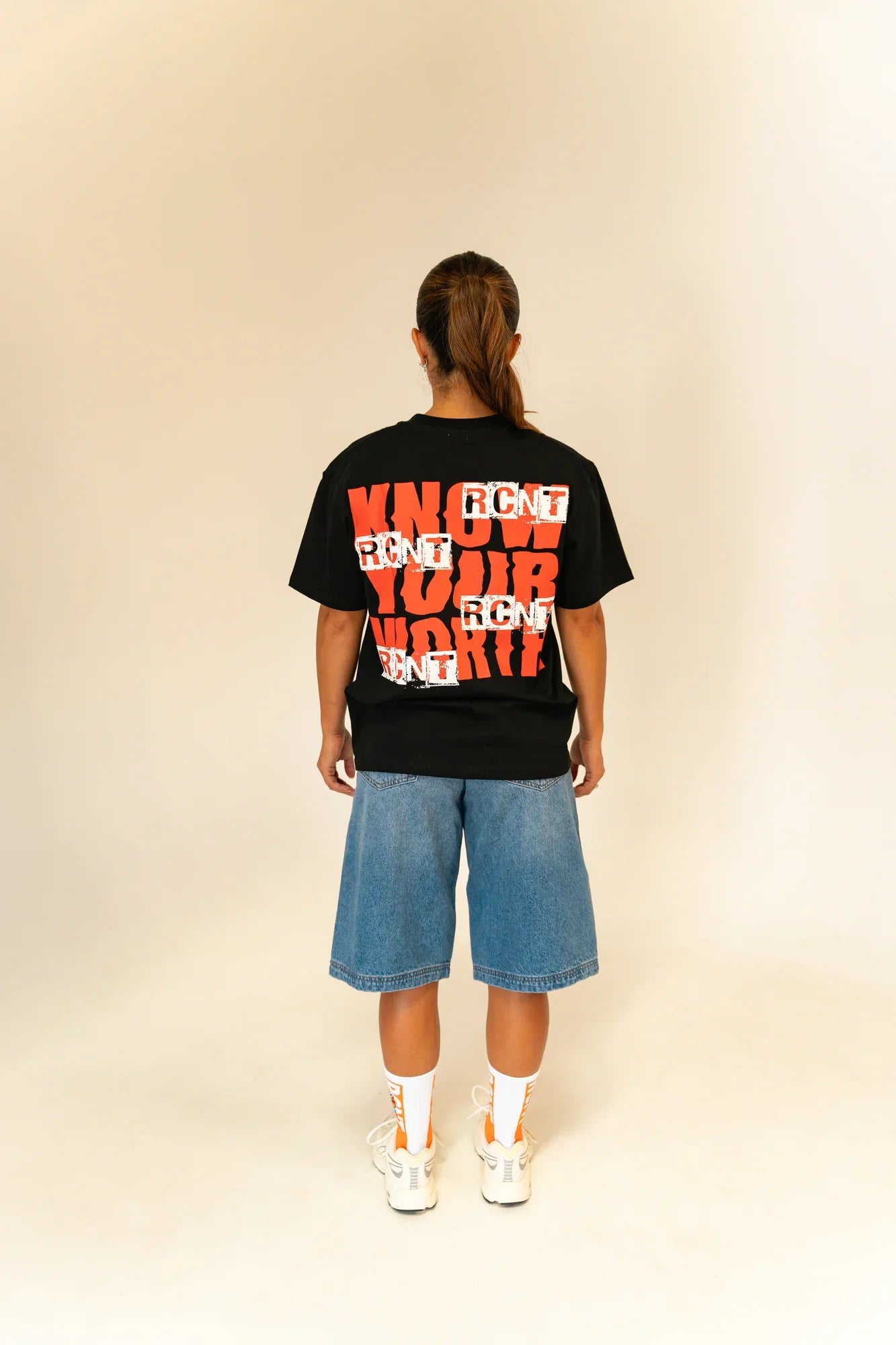 Back view of person wearing black 'Know Your Worth' graphic tee, denim shorts, and white sneakers with socks