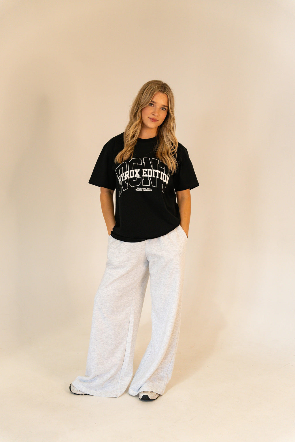 Young woman in black Hyrox Edition t-shirt and light gray wide-leg pants standing against beige background