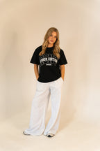 Young woman in black Hyrox Edition t-shirt and light gray wide-leg pants standing against beige background