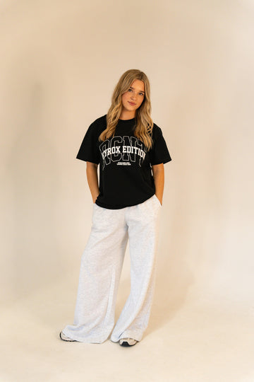 Young woman in black Hyrox Edition t-shirt and light gray wide-leg pants standing against beige background
