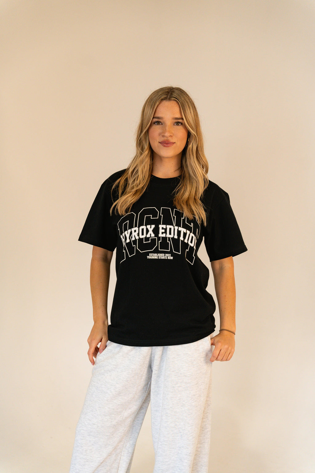 Woman wearing black ICON Hyrox Edition T-shirt and light gray sweatpants against beige background