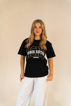 Woman wearing black ICON Hyrox Edition T-shirt and light gray sweatpants against beige background