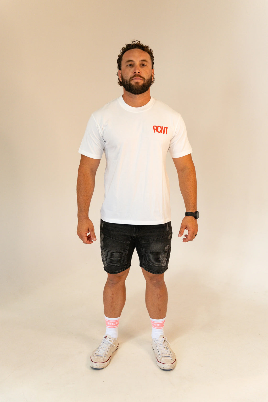 Man wearing white RCNT logo t-shirt, black distressed shorts, white RCNT socks, and white sneakers on plain background